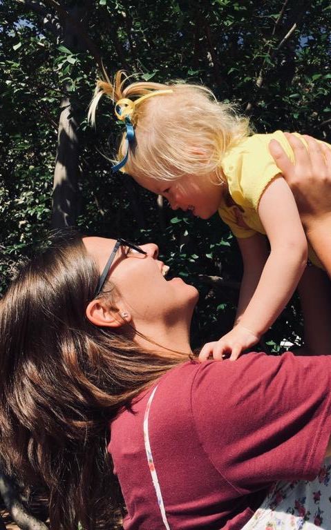 A parent lifting a small child into the air, the two of them laughing face to face under a tree